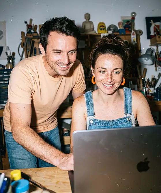 Un homme et une femme souriants regardent un ordinateur portable dans un atelier avec des outils en arrière-plan.