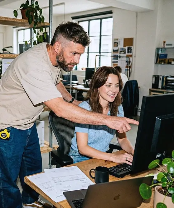 Un homme montre quelque chose sur un écran d'ordinateur à une femme souriante assise à un bureau dans un bureau moderne.
