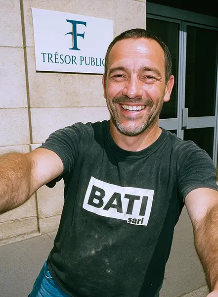 Artisan maçon souriant en t-shirt BATI sarl prenant un selfie devant un panneau Trésor Public.
