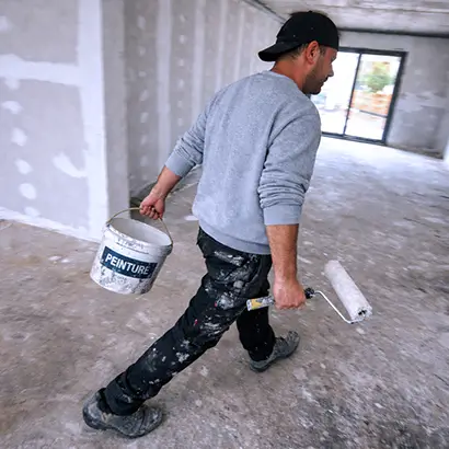 Peintre en bâtiment marche dans une pièce avec un pot de peinture et un rouleau à peinture.