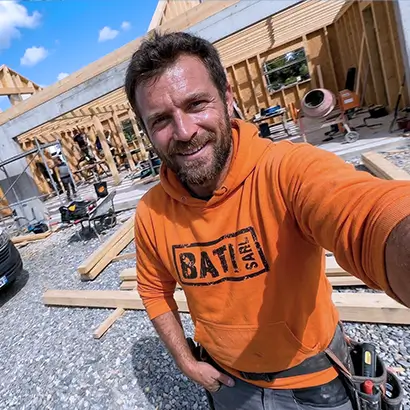 Charpentier souriant en sweat à capuche orange sur un chantier de construction en bois.