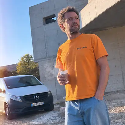 Un plombier en t-shirt orange tenant un gobelet sur un chantier avec un fourgon blanc Mercedes en arrière-plan.
