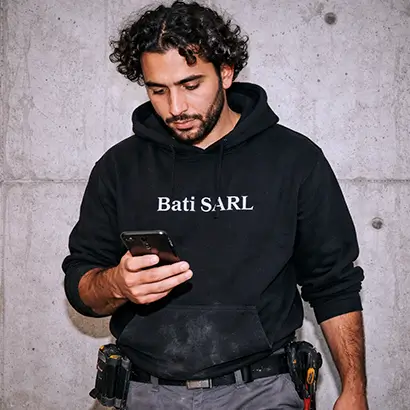 plaquiste platrier en sweat-shirt Bati SARL regarde son téléphone sur un chantier.