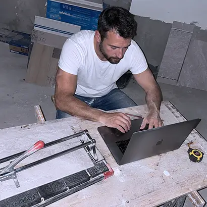 Carreleur tape sur un ordinateur portable sur un chantier avec des outils de carrelage à proximité.