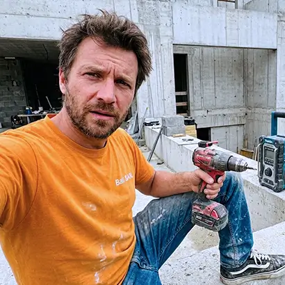 Couvreur en T-shirt orange tenant une perceuse sur un chantier de construction en béton.