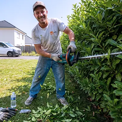 Paysagiste souriant taille une haie avec un taille-haie électrique dans un jardin ensoleillé.