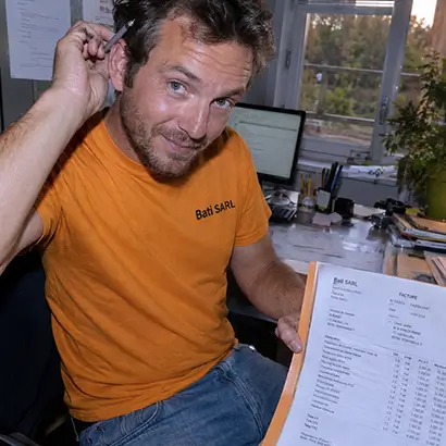 Artisan souriant montrant une facture dans un bureau avec ordinateur et fenêtres ouvertes.