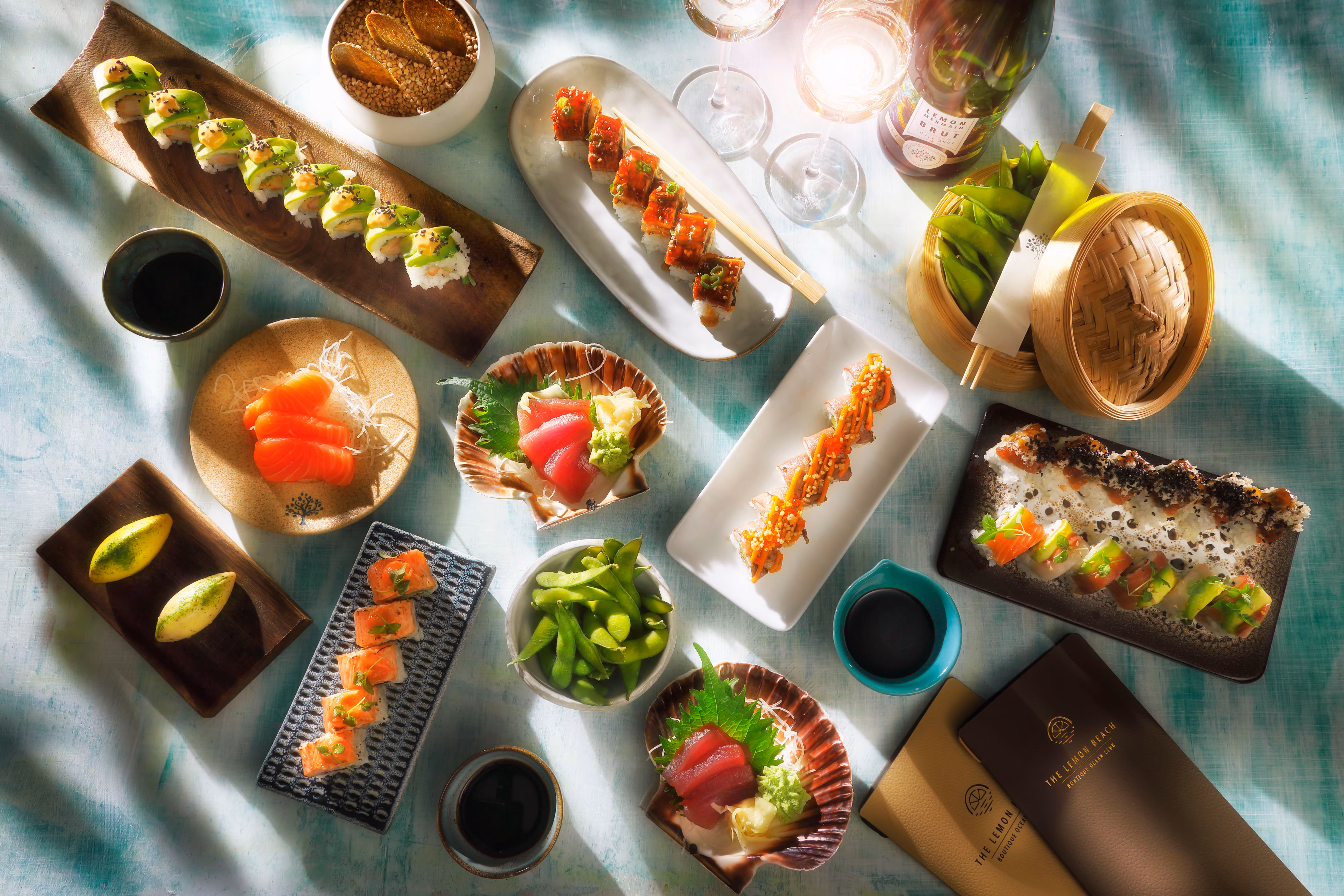 Assorted sushi and sashimi dishes with edamame, soy sauce, and a bottle of sparkling wine arranged on a light blue tablecloth.