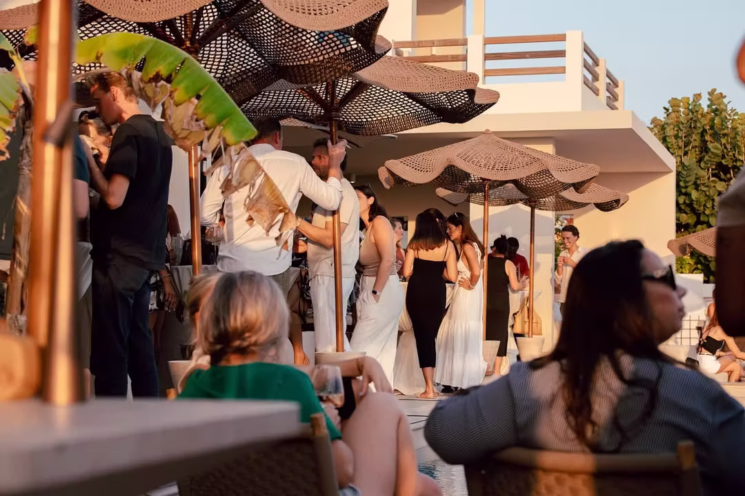People socializing under woven patio umbrellas at an outdoor gathering in warm sunlight.