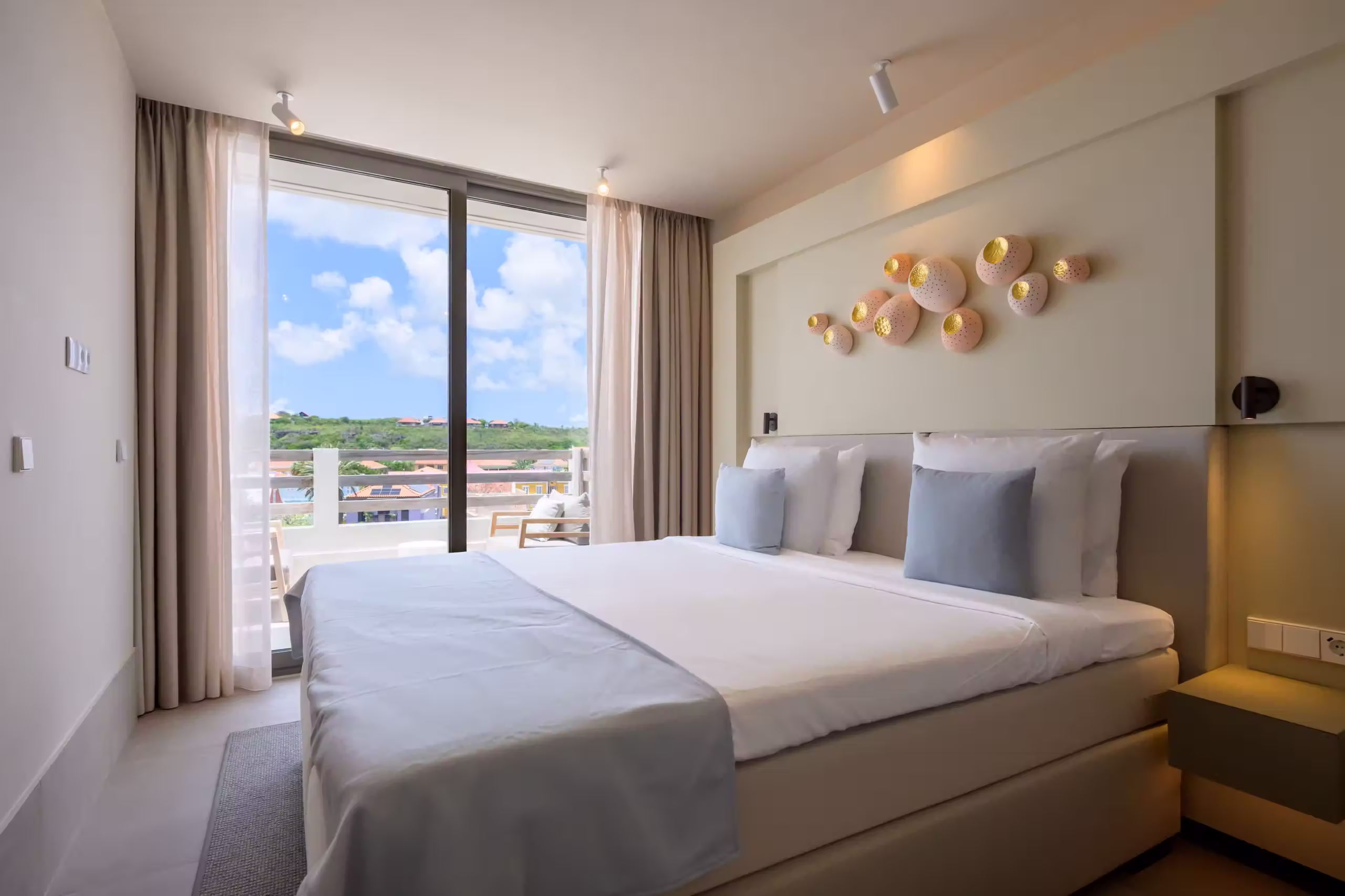 Modern hotel bedroom with a large bed, light blue pillows, decorative wall art, and a balcony with patio chairs overlooking a green landscape under a blue sky.