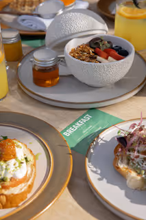 Breakfast spread featuring a bowl of granola with berries, honey jar, orange juice, and two plates with savory dishes on a table.