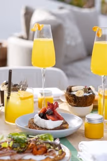 Table with brunch items including yellow cocktails with orange garnish, a plate with toast topped with cream and berries, a coconut bowl with bananas, and jars of yellow juice.