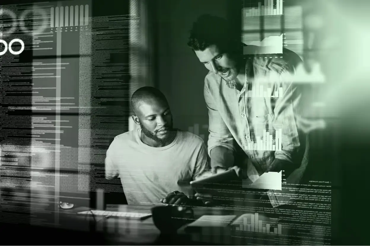Two men collaborate over a tablet at a desk, one seated and one leaning in, amid translucent charts and data overlays, creating a focused, analytical mood.