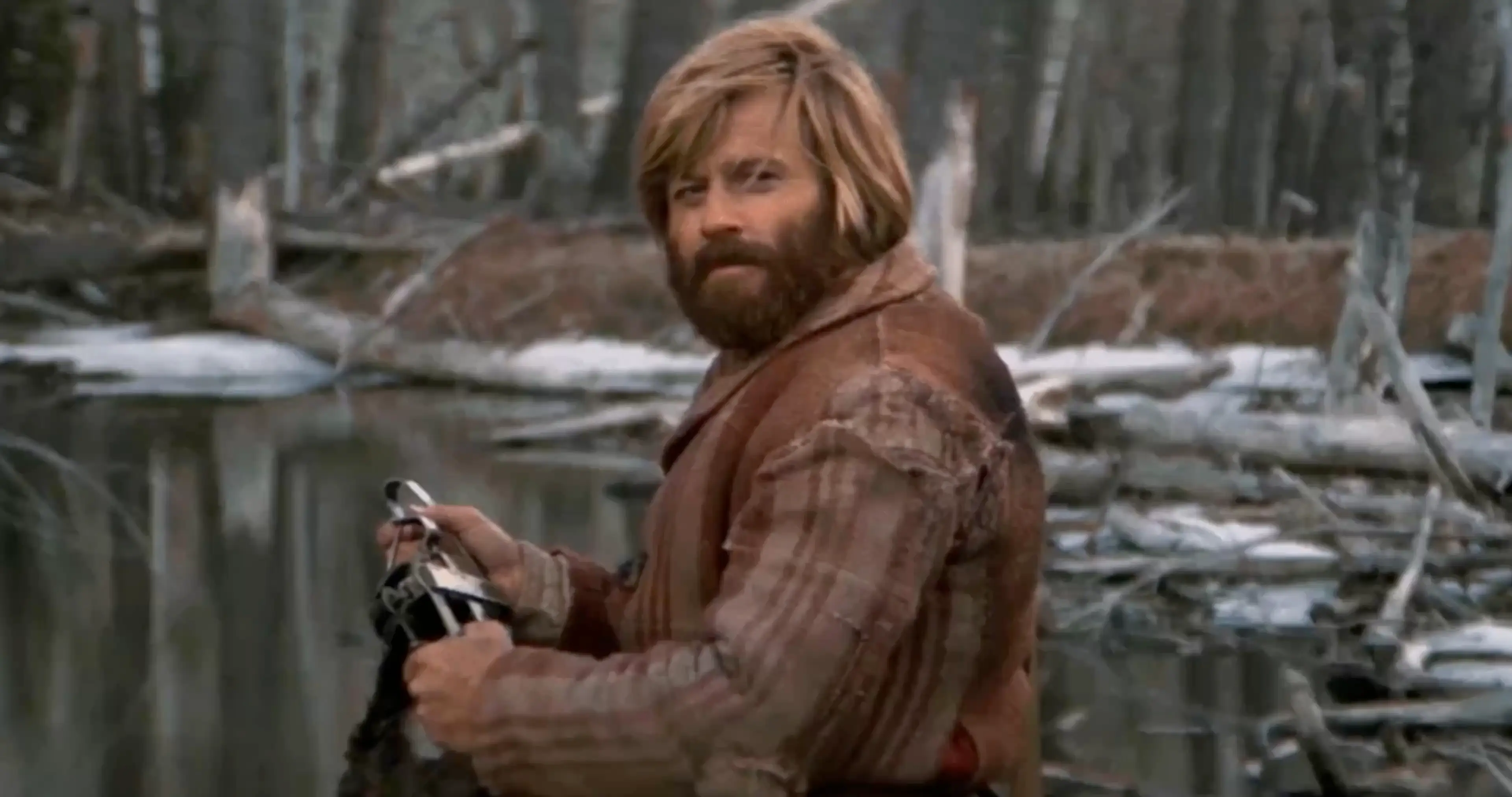 Bearded man with long hair wearing a worn brown coat stands near a body of water holding an object, with a forest background.