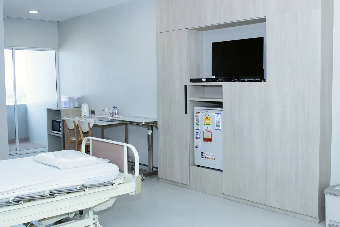 Minimalist patient room with a bed, TV, fridge, and small dining area.