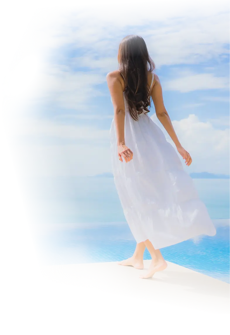 Woman in a flowing white dress walking barefoot by an infinity pool, facing the ocean under a bright blue sky.