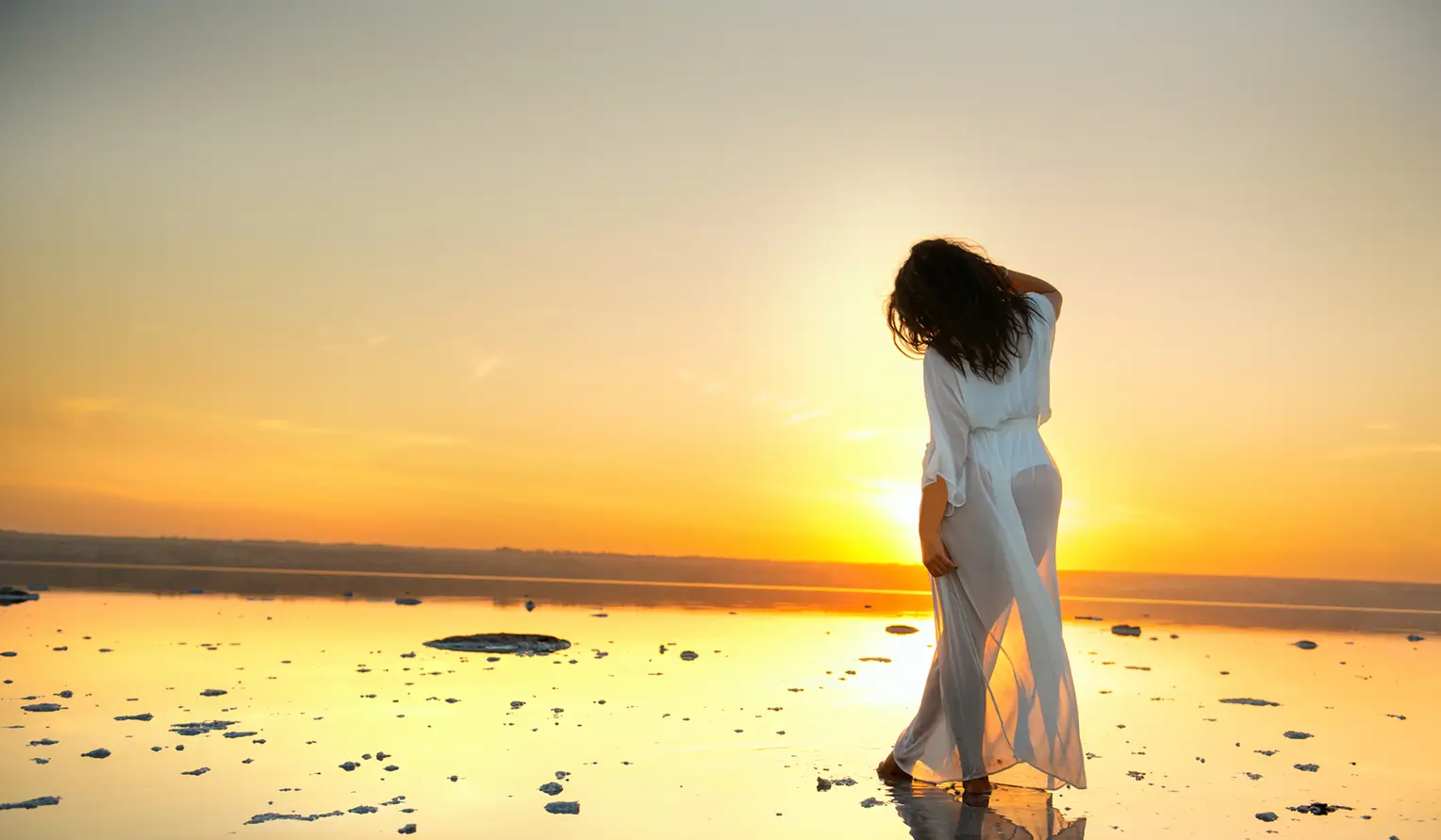 Woman in a flowing white dress standing on a reflective salt flat at sunset, gazing into the horizon.