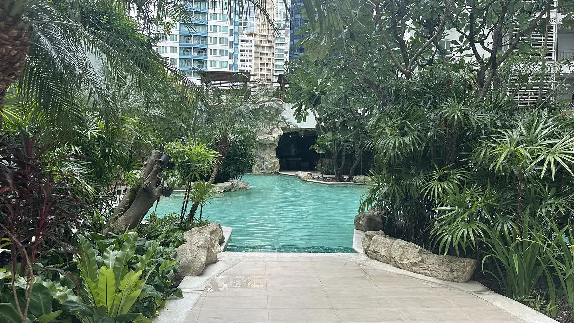 Tropical-style outdoor pool surrounded by greenery and tall buildings.