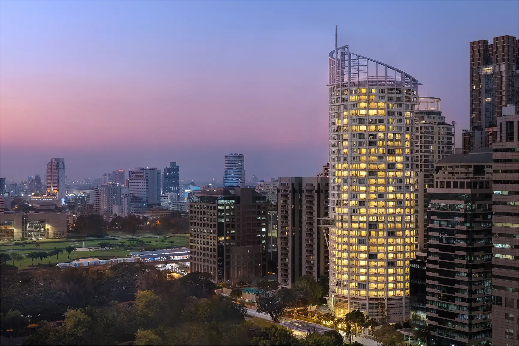 Dusit Suites Hotel exterior lit up at dusk in Bangkok cityscape.