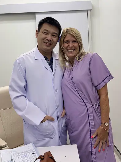 A doctor and a woman wearing purple clinic attire smile together in a medical office.