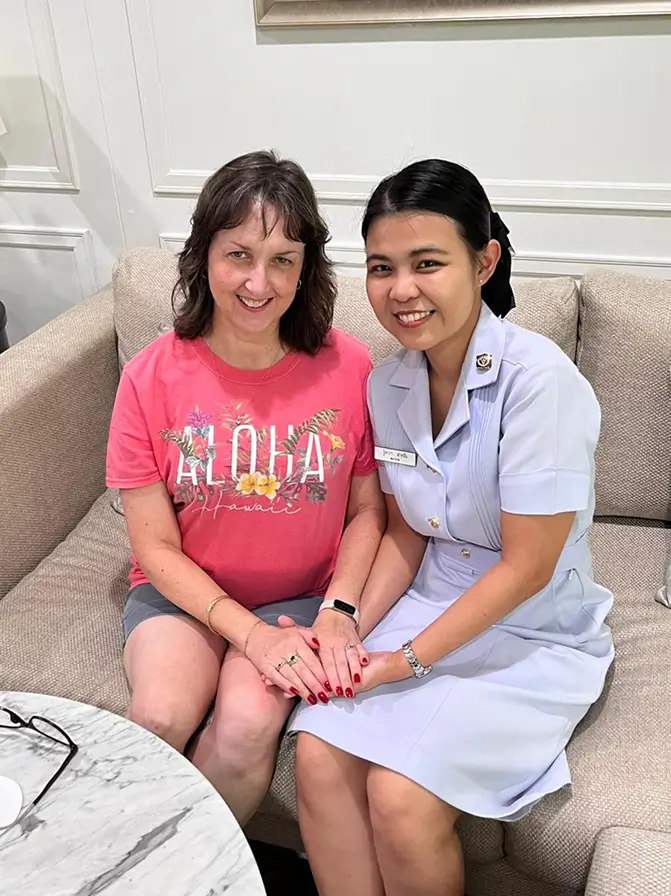 Two women sitting on a beige couch holding hands and smiling, one wearing a pink 'Aloha Hawaii' T-shirt and the other in a light blue uniform dress.