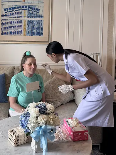 A nurse in a purple uniform tends to a seated woman in a light blue shirt inside a hotel suite.