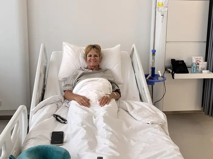 A woman smiles while lying in a hospital bed with medical equipment nearby.
