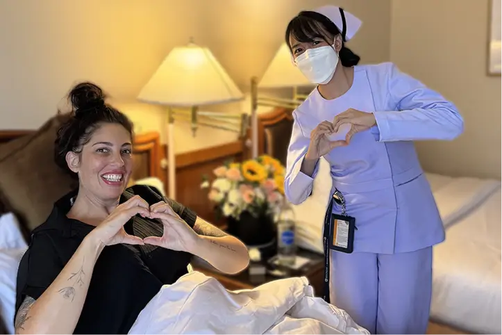 Patient in bed forming a heart shape with her hands beside smiling nurses.
