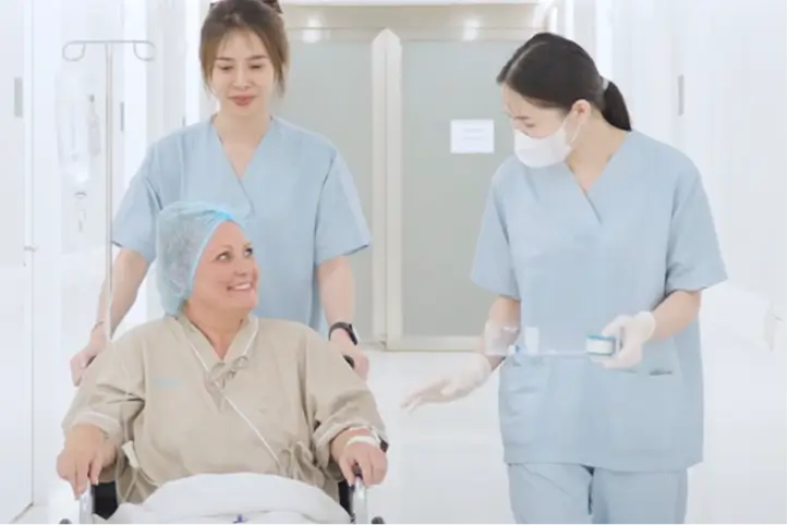 Two nurses in scrubs assisting a patient in a wheelchair along a hospital hallway.
