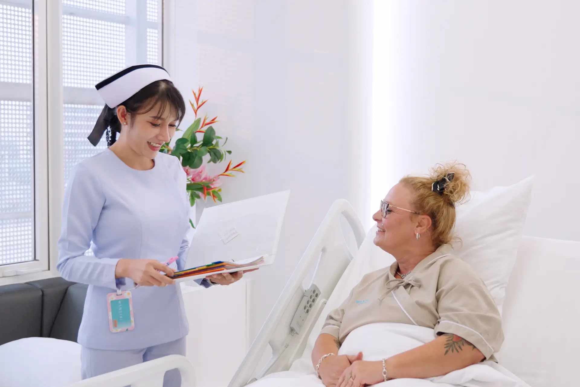 A nurse smiling while holding a folder and talking to a female patient sitting up in a hospital bed.