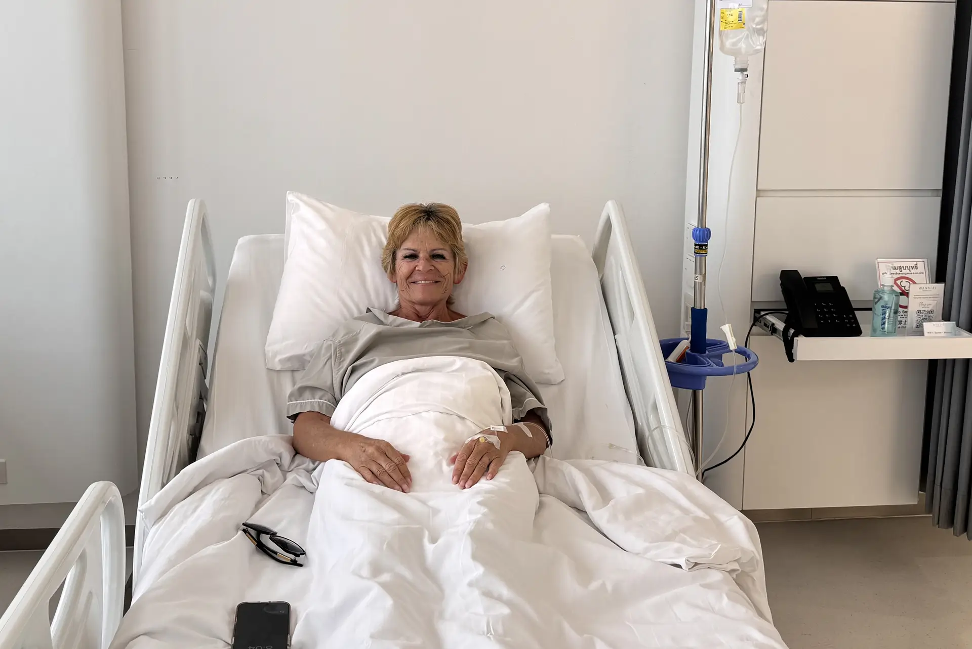 Smiling woman lying in a hospital bed with IV drip attached, covered with a white blanket.