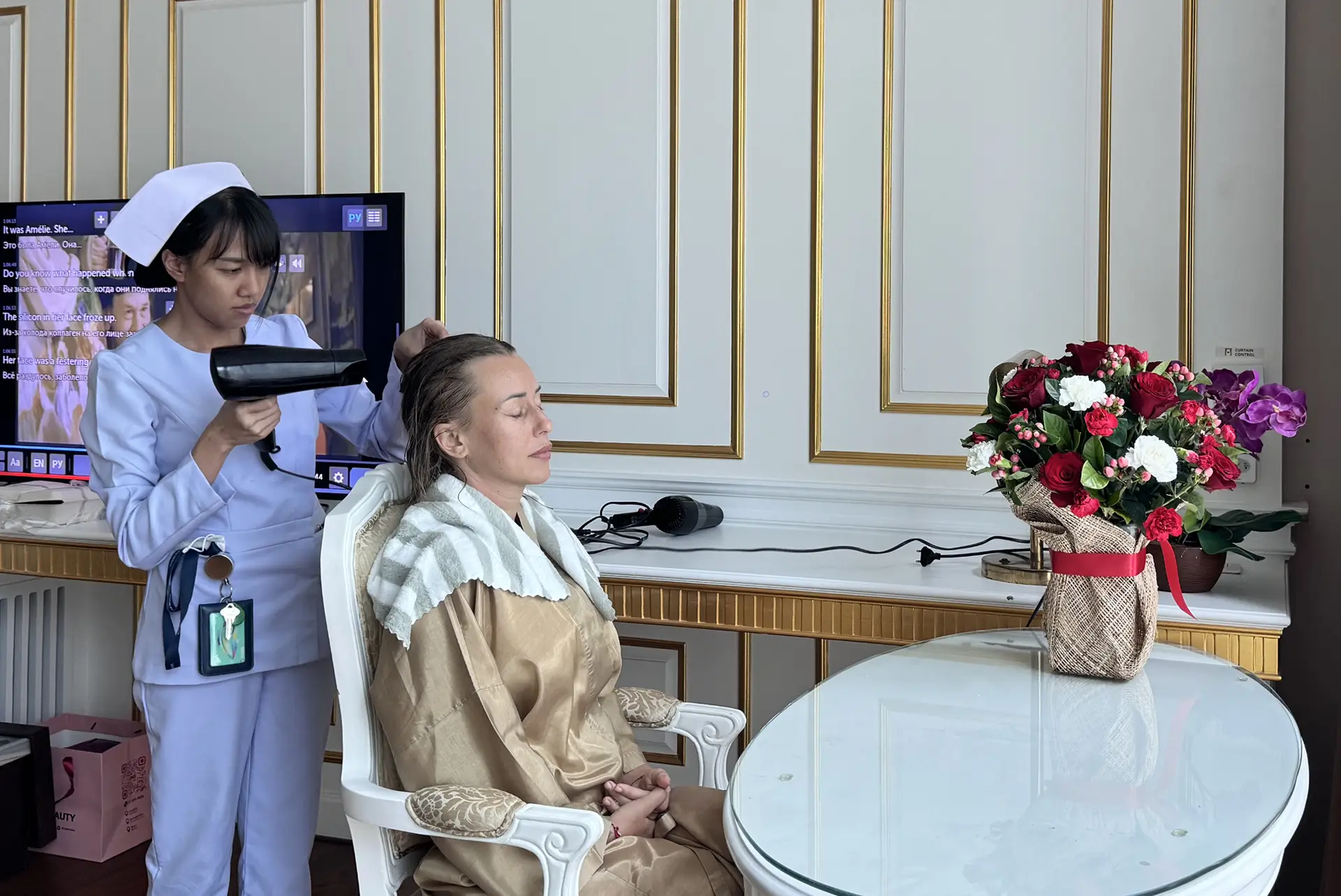 A nurse in a light blue uniform and cap blow-dries a seated woman's wet hair in a softly lit room with gold-trimmed white walls and a vase of red and white flowers on a glass table.