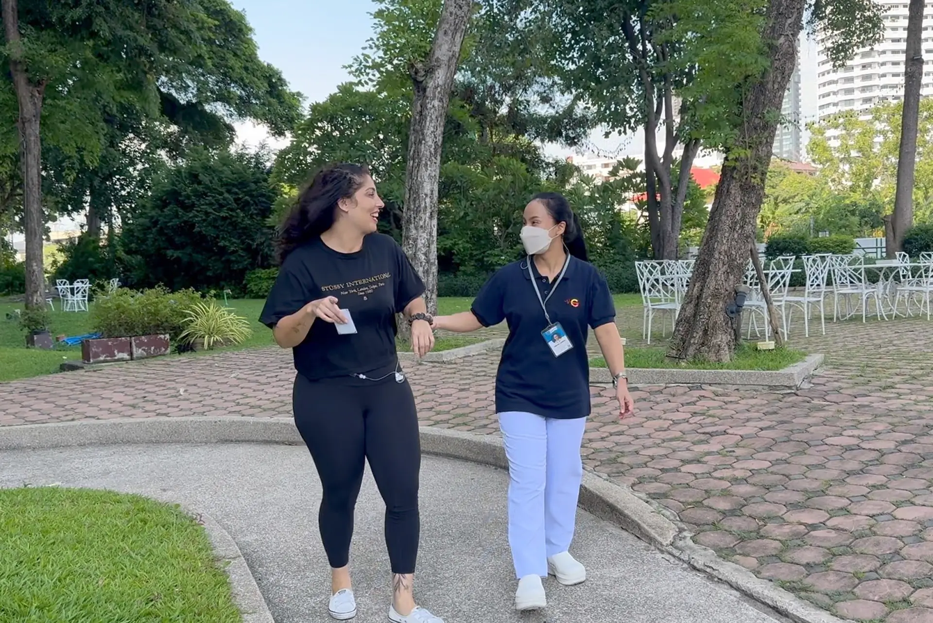 A woman in black athletic wear walking outdoors with a masked healthcare worker in blue uniform holding her arm.