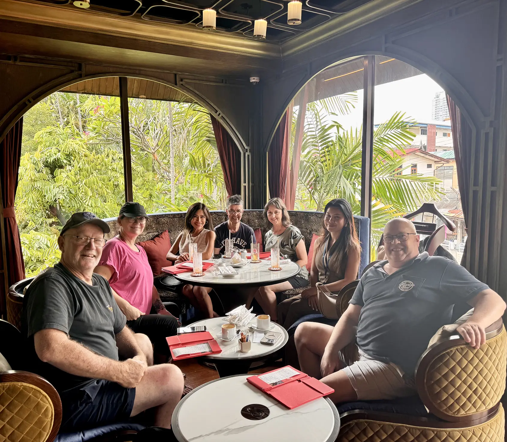 Group of seven people sitting around two circular marble tables in a cozy cafe with large arched windows and green plants outside.