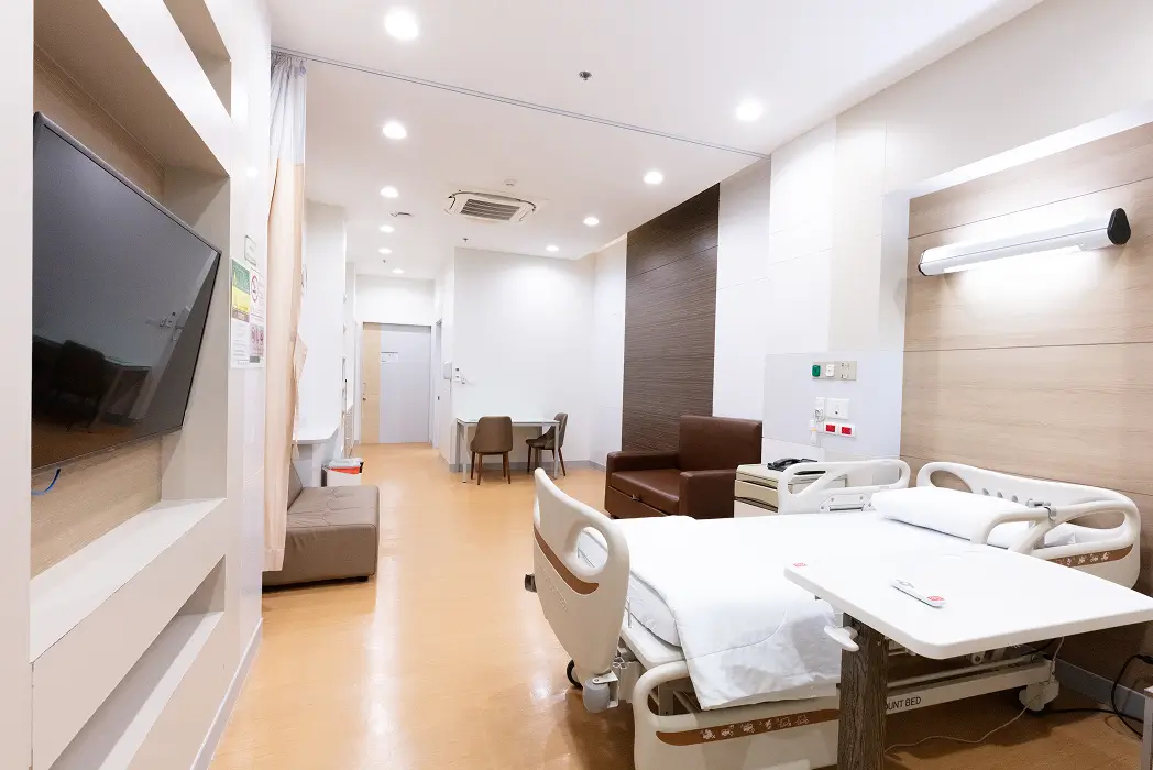 Bright hospital room with a patient bed, brown armchair, two chairs with a table, wall-mounted TV, and ceiling lights.