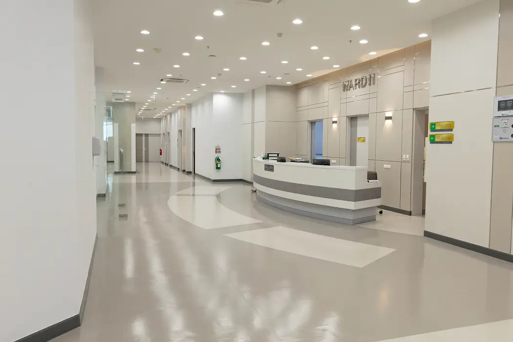 Bright, empty hospital corridor with a curved reception desk labeled Ward II and multiple doors along the hallway.