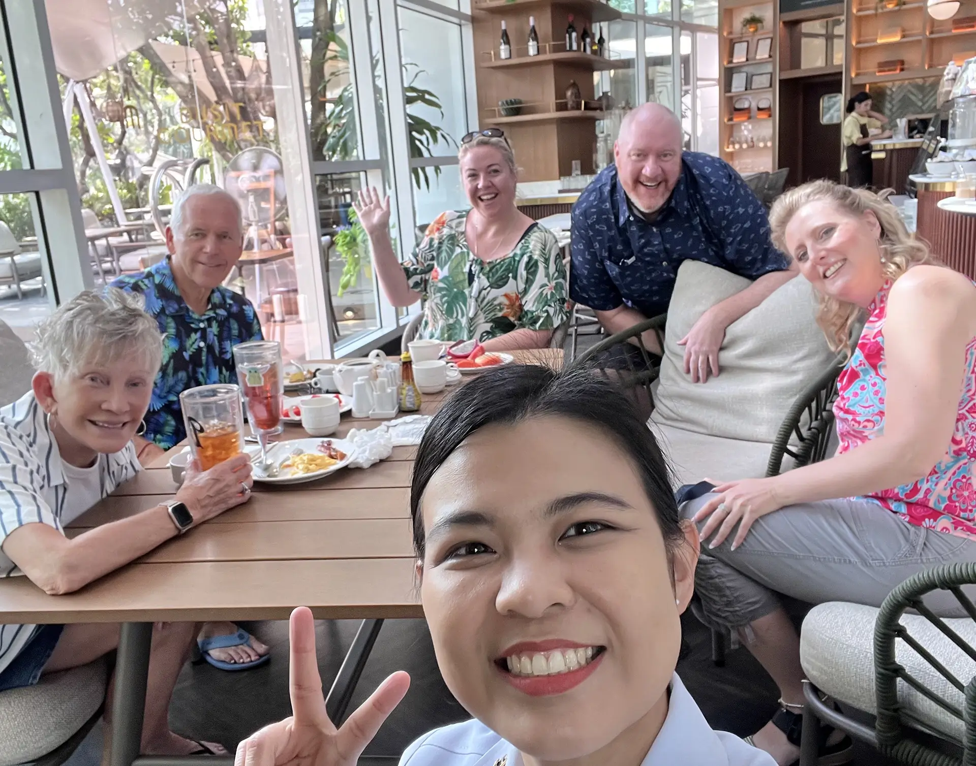 Group of six adults smiling and posing for a selfie around a table with breakfast plates and drinks in a bright café.
