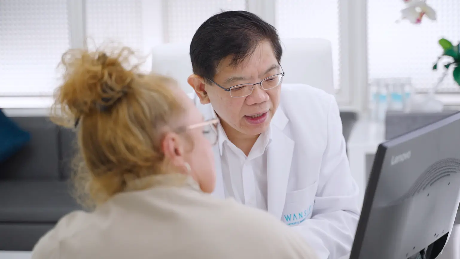 Doctor in white coat discussing something with a patient while looking at a computer screen.