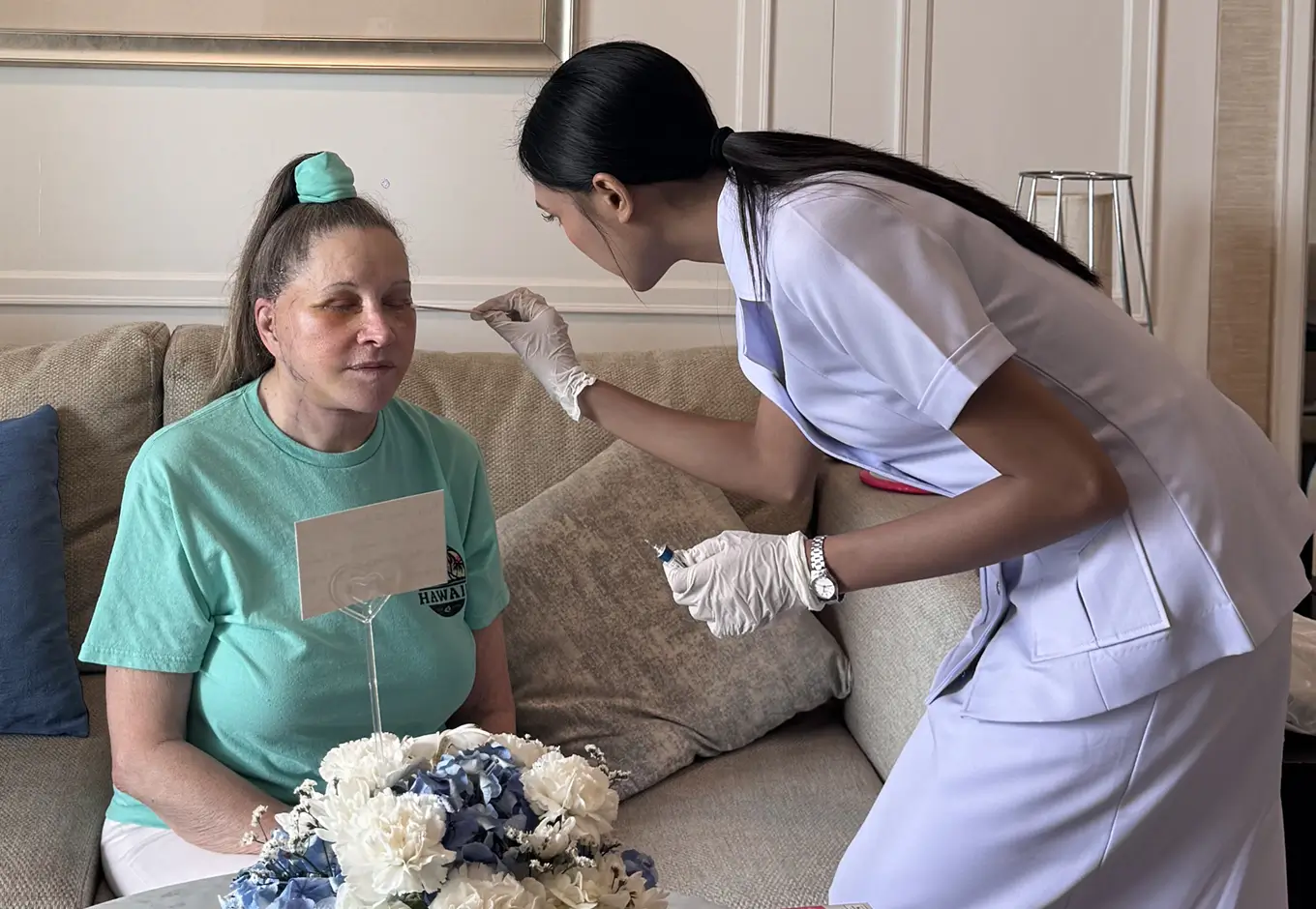 Caregiver in white uniform and gloves administering a nasal swab test to a seated woman in a green shirt on a beige sofa.