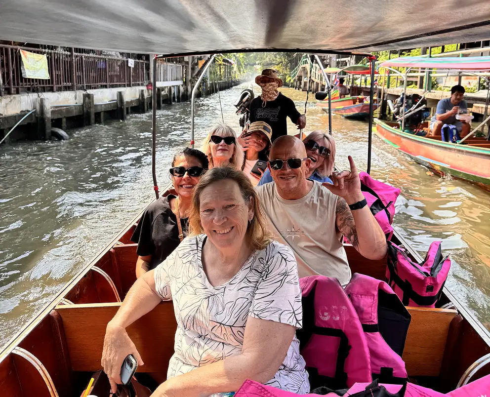 Group of people smiling and sitting in a covered wooden boat on a canal, with pink life jackets and another boat nearby.