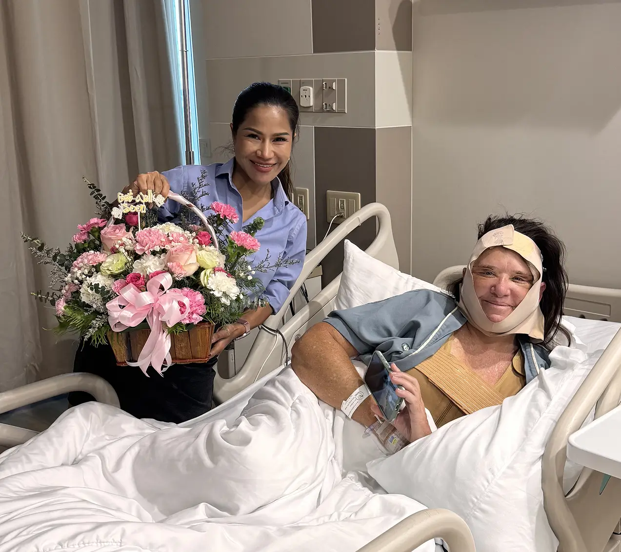 Smiling woman holding a basket of pink and white flowers beside a hospital bed where a patient with bandages on her face is lying and holding a phone.