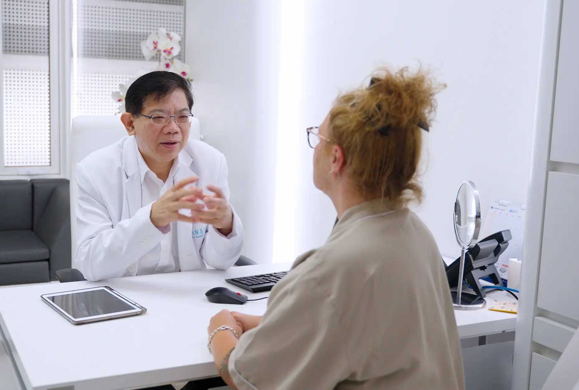 Male doctor in a white coat talking to a female patient across a desk in a bright medical office.