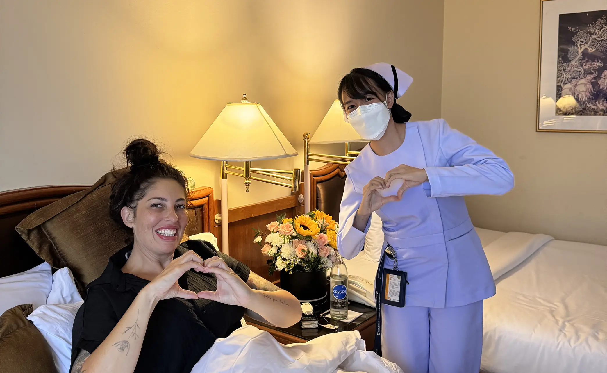 A smiling woman lying in bed and a nurse wearing a mask, both forming heart shapes with their hands in a warmly lit room.