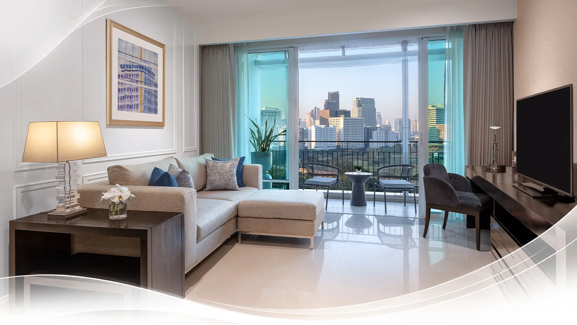 Modern living room with beige sofa, side table lamp, balcony overlooking city skyline, and TV on desk.