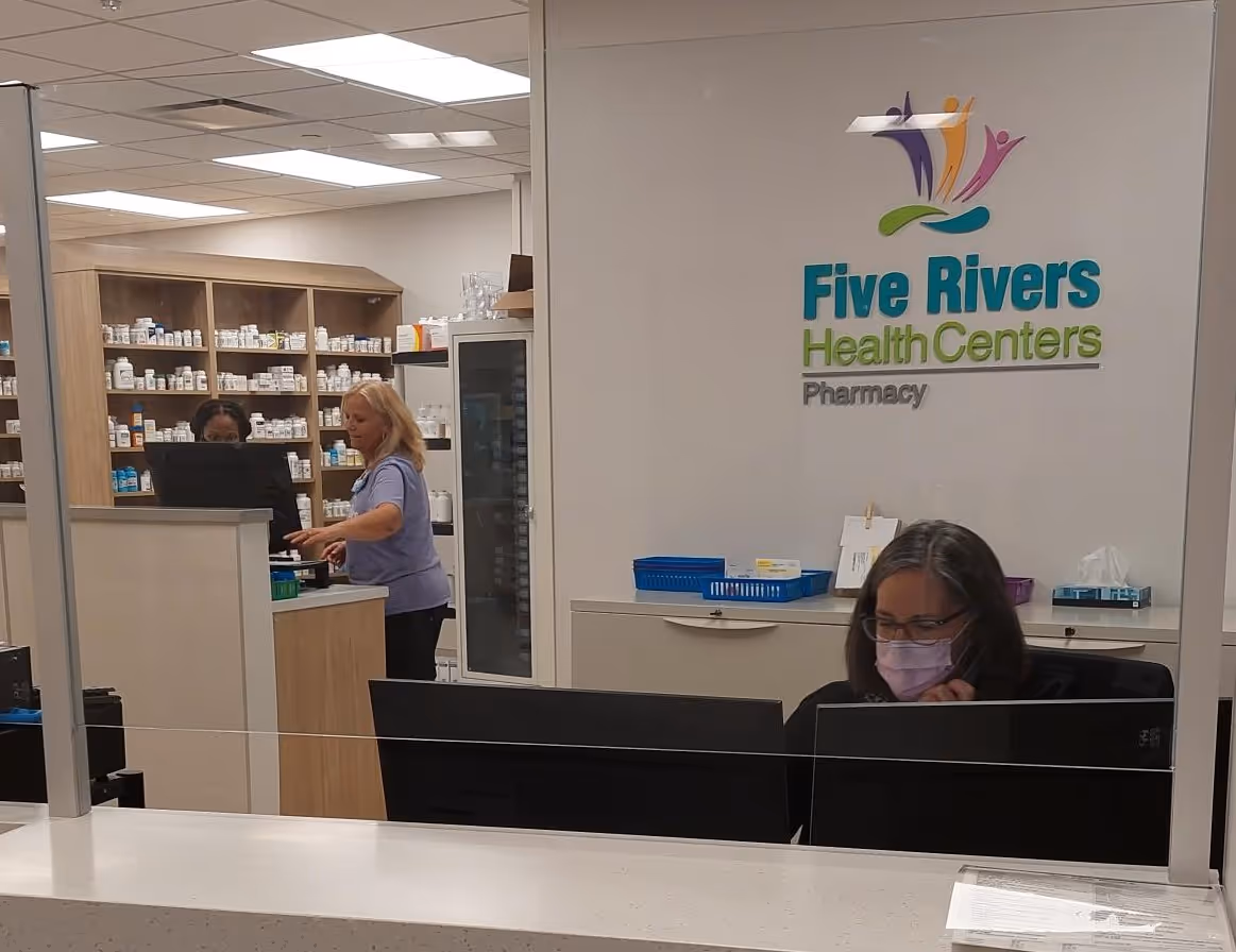 Pharmacy counter at Five Rivers Health Centers with staff working behind computer screens and shelves stocked with medication bottles in the background.