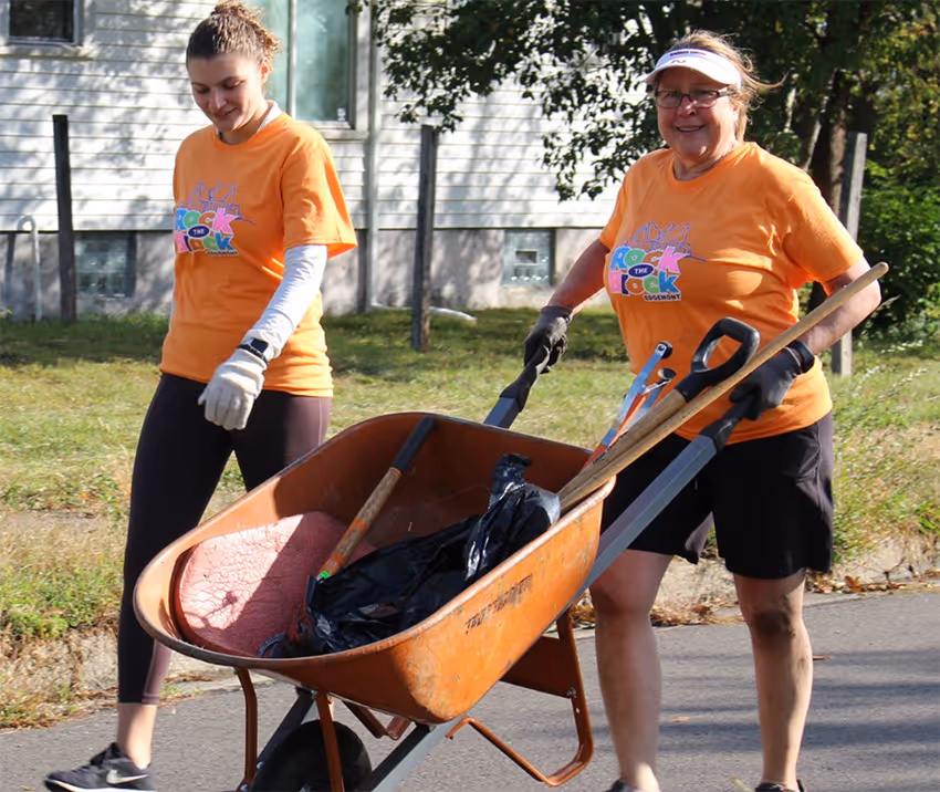 Two women wearing orange Rock the Block Edgemont t-shirts, one pushing an orange wheelbarrow filled with tools and bags.