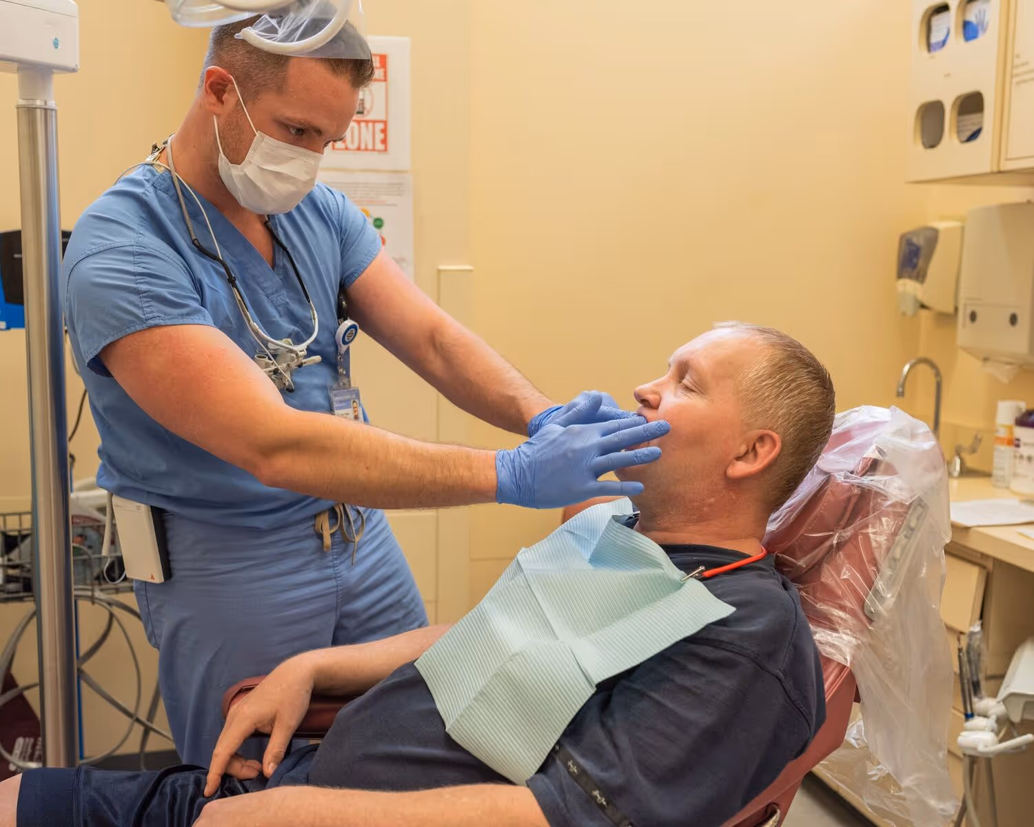 Dentist wearing blue scrubs and gloves examining a patient's mouth in a dental office.