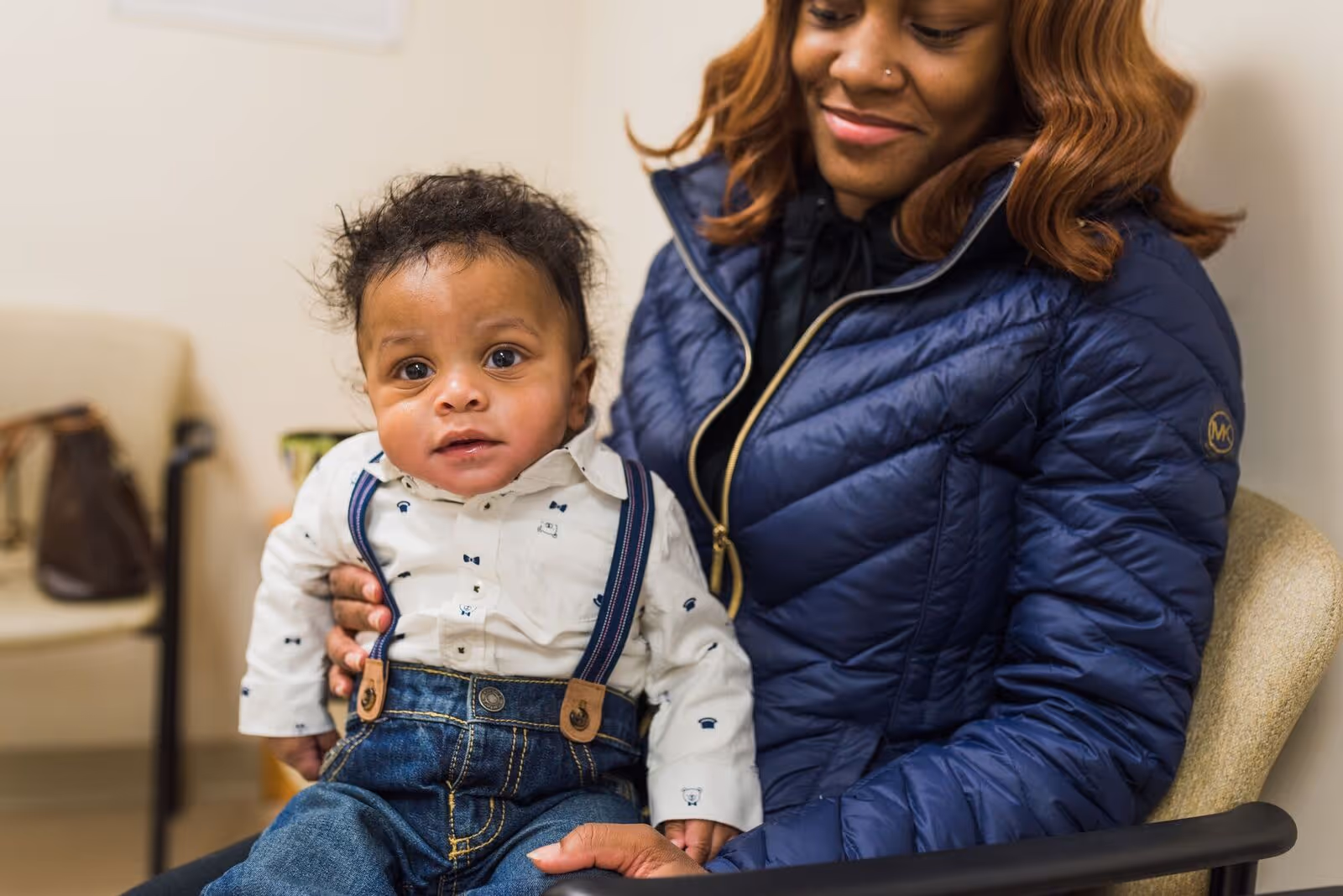 Smiling woman in a blue jacket holding a baby dressed in a white shirt with suspenders and denim pants.