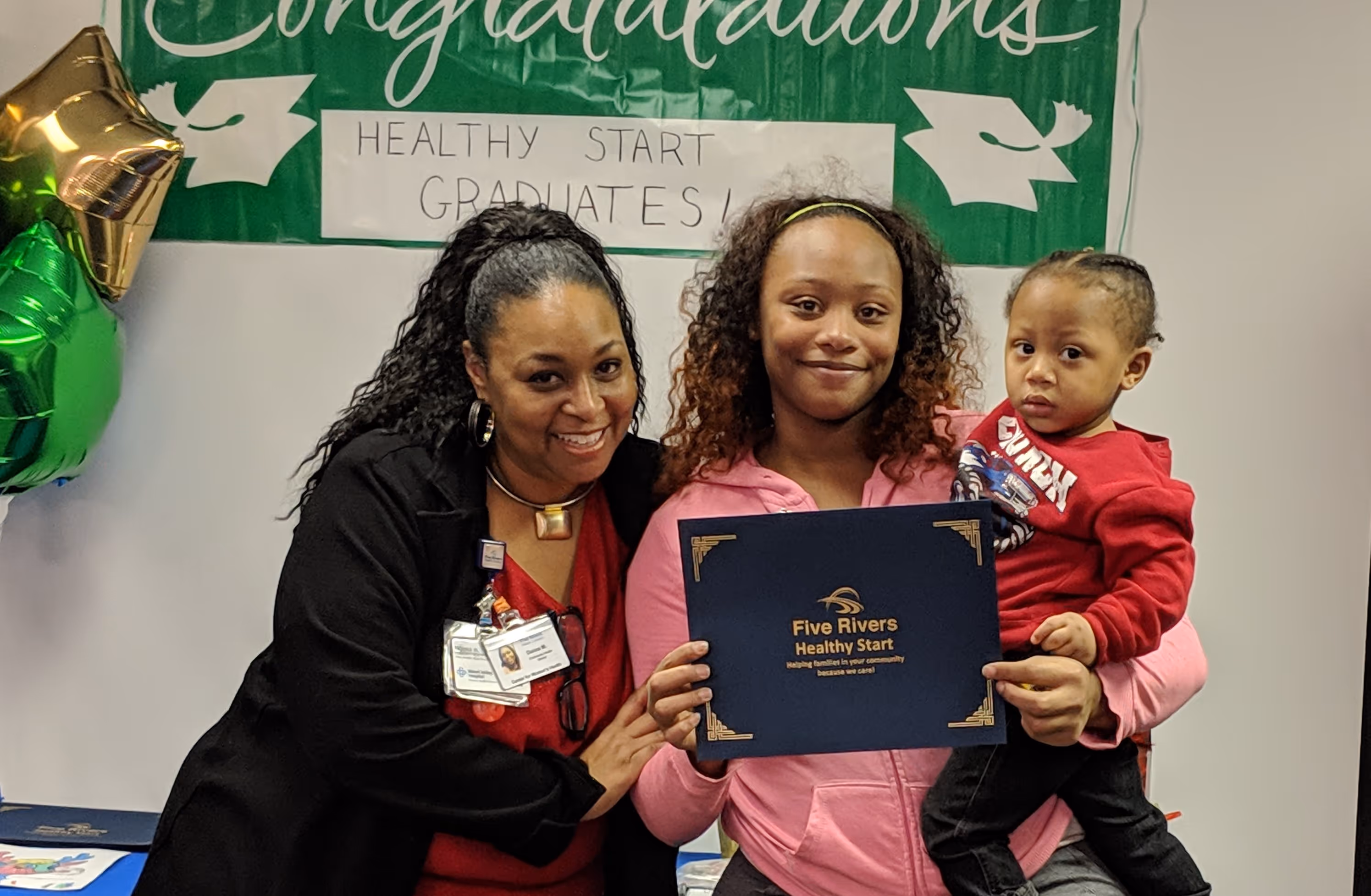 Two women and a child posing with a Five Rivers Healthy Start certificate in front of a 'Healthy Start Graduates' banner with green and gold balloons.