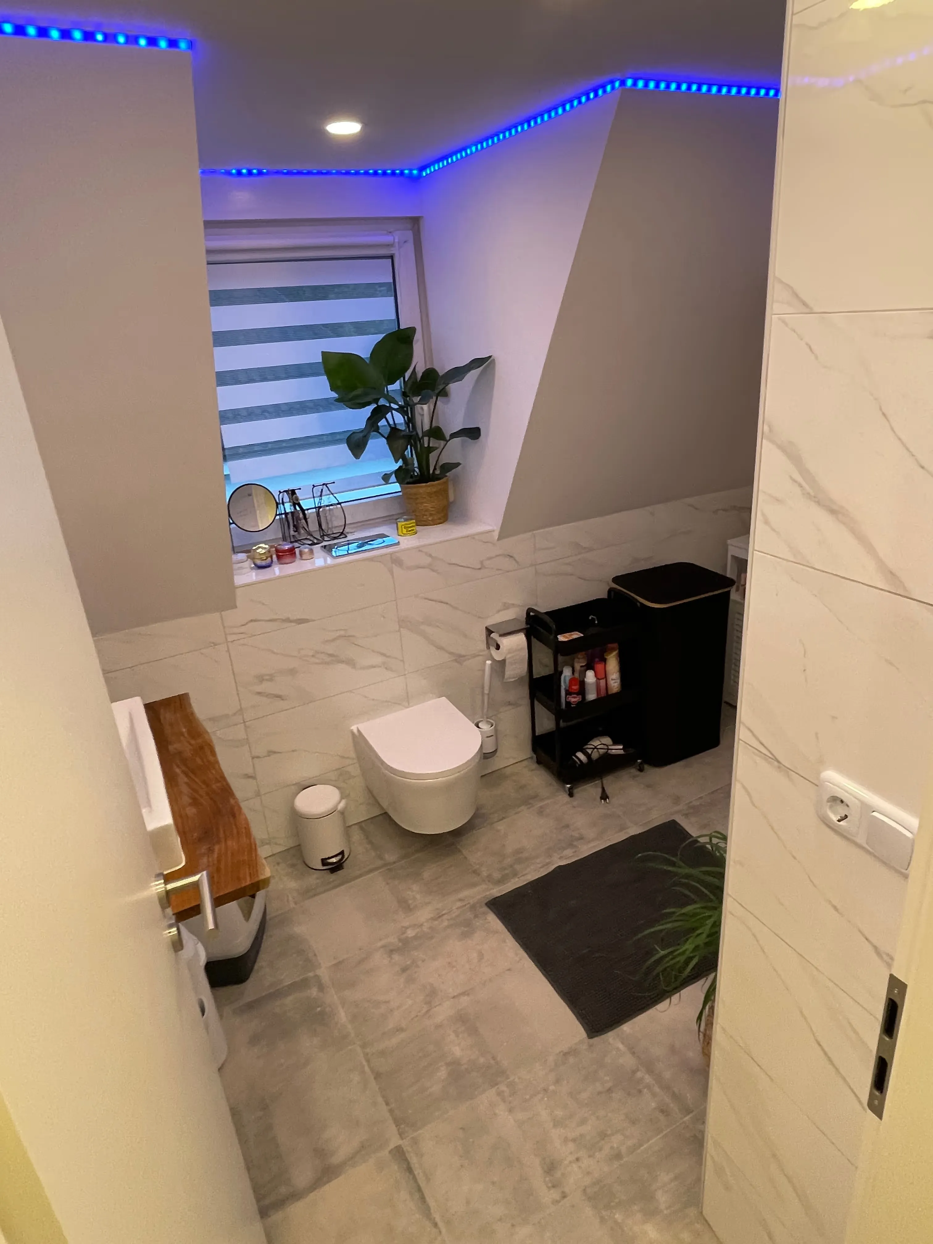 Modernes Badezimmer mit weißen Fliesen, wandhängendem WC, Holzregal, Pflanzen und blauen LED-Leisten an der Decke.